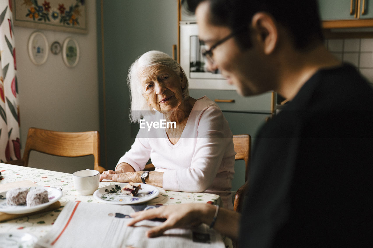 Young male nurse reading newspaper while elderly woman looking at him in kitchen