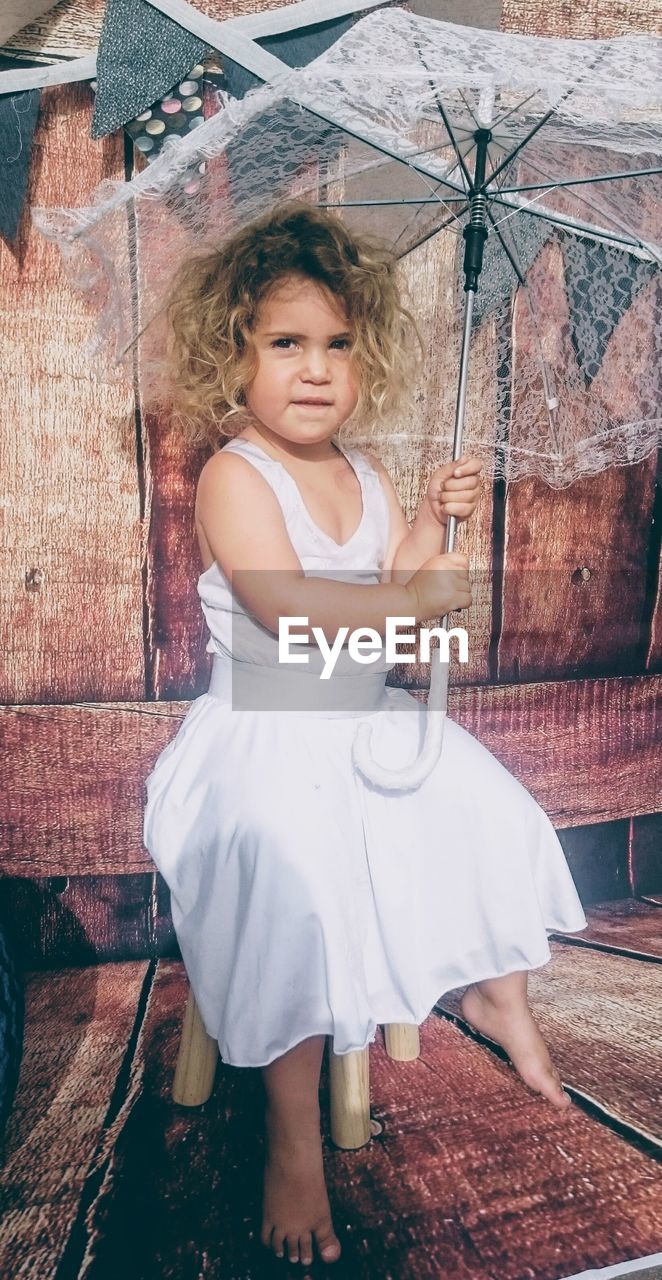 Portrait of girl holding umbrella sitting on stool against wall