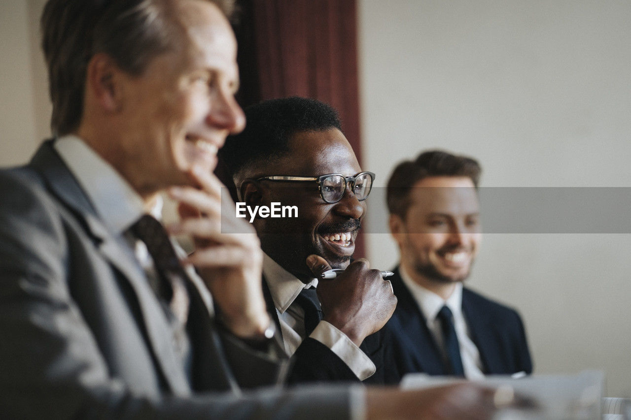 Happy mature businessman with hand on chin by male colleagues in meeting at office