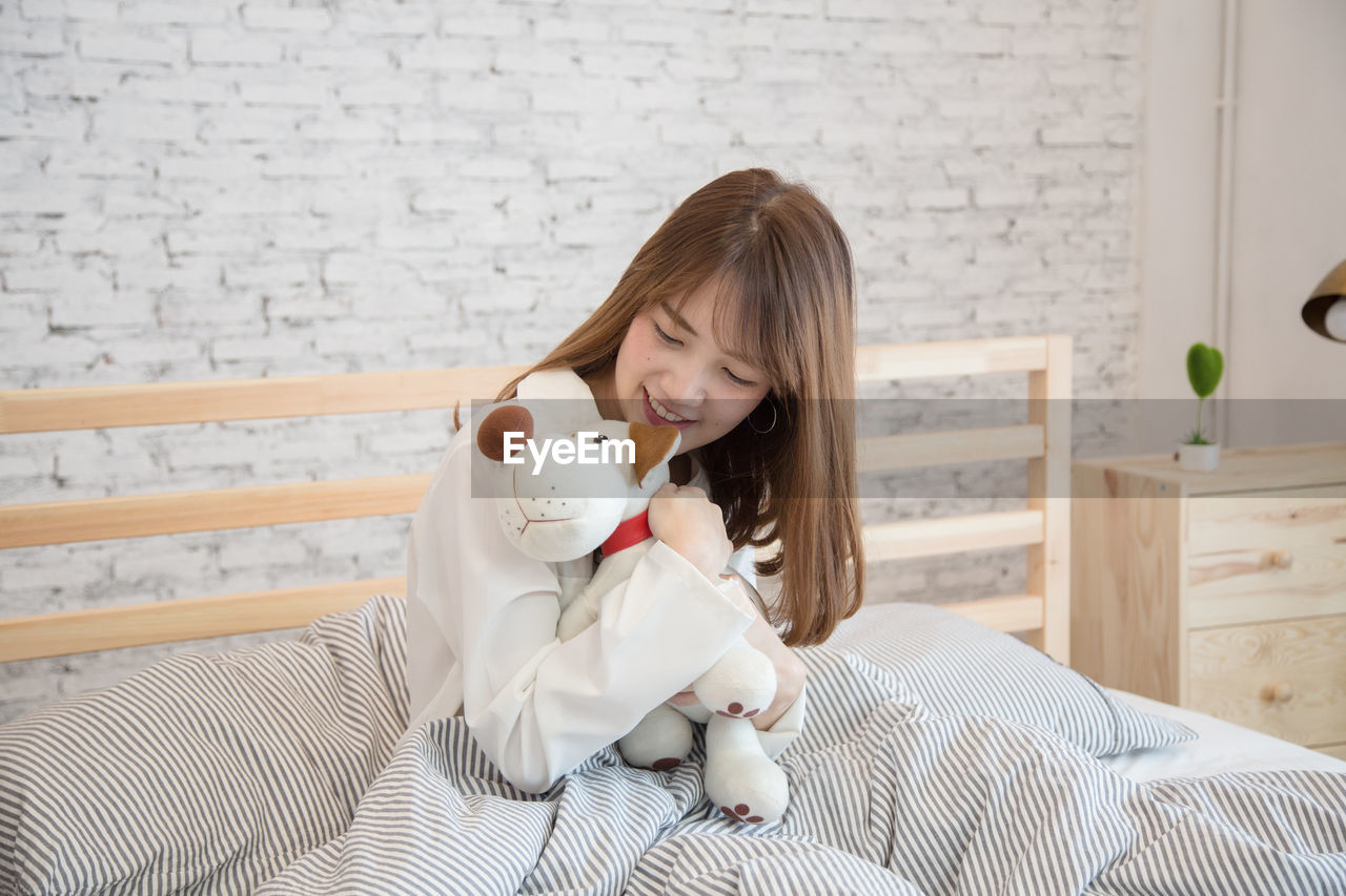 Smiling young woman embracing toy while sitting on bed at home