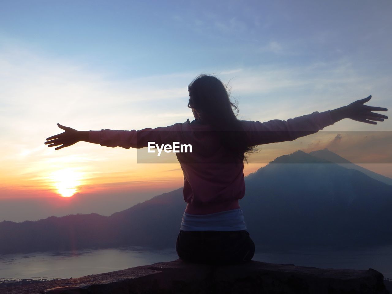 Rear view of woman with arms outstretched while sitting on rock against sky during sunset