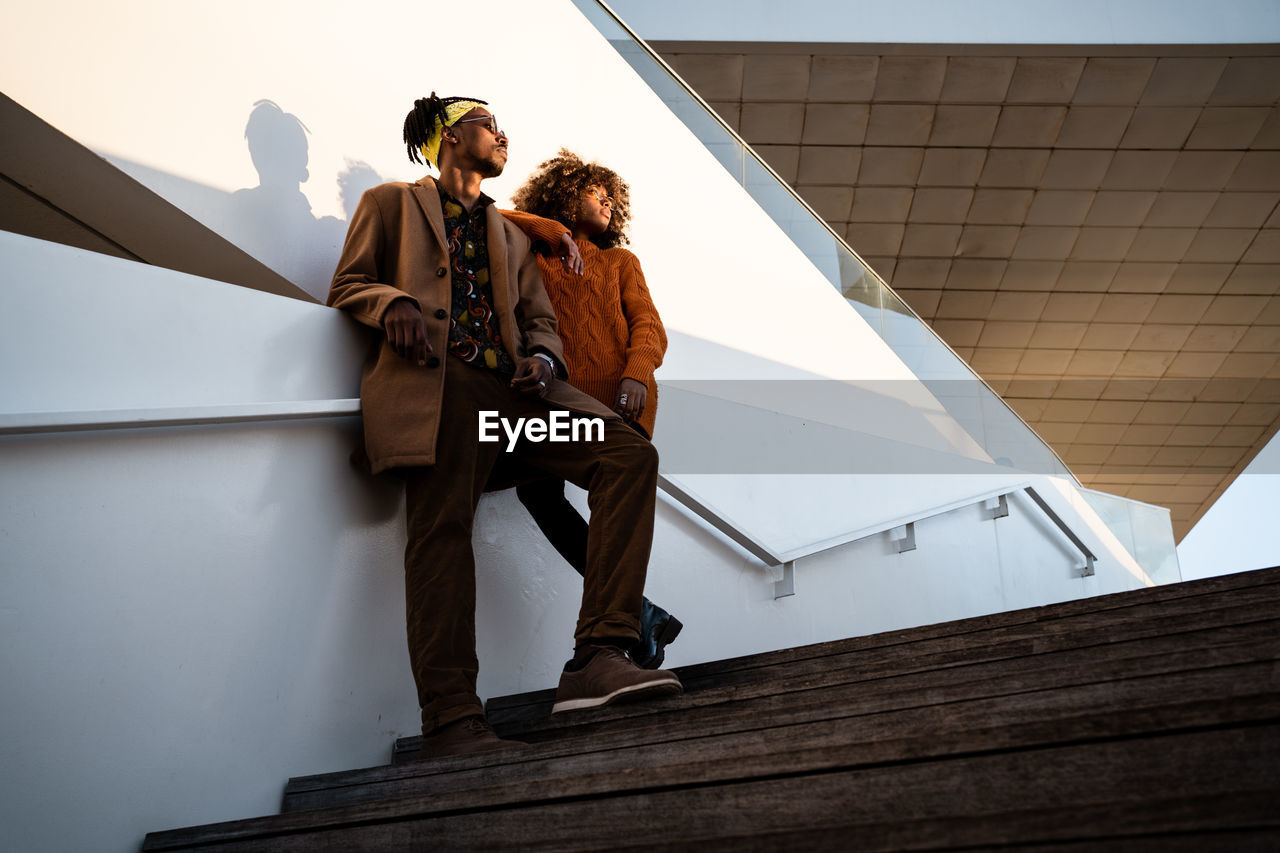 From below of cool african american woman and man in trendy clothes standing together on stairs in sunset light looking away