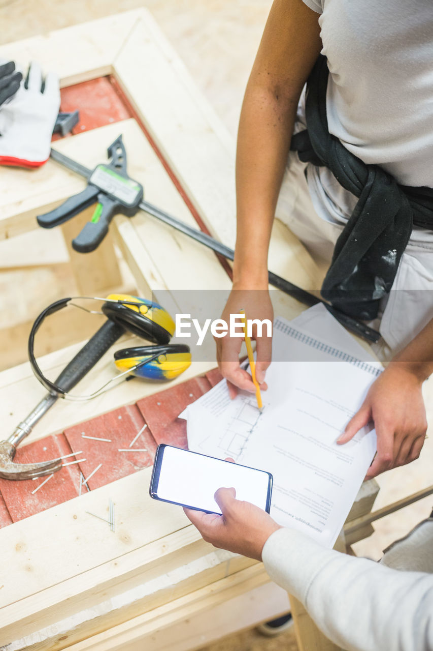 Teenage trainee holding smart phone by coworker drawing diagram on paper at workshop