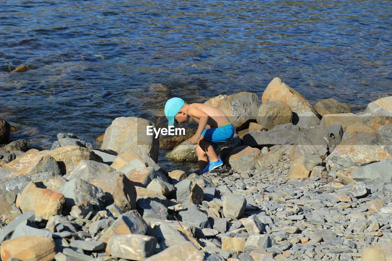 Four-year-old boy in cap, blue bathing shorts and rubber slippers enters the sea on the rocks