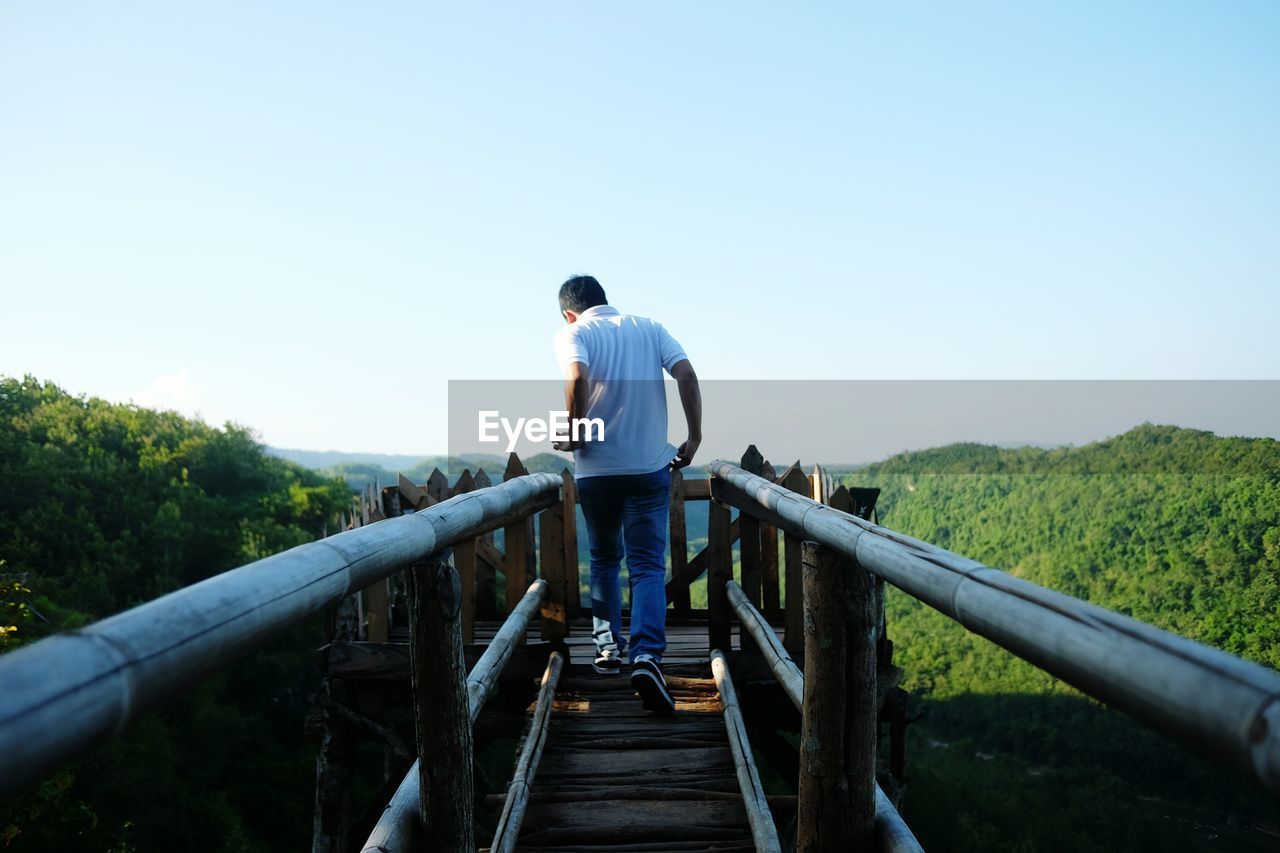 Rear view of man walking on footbridge against clear sky