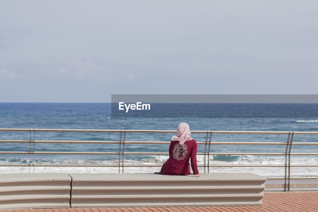 WOMAN SITTING ON RAILING AGAINST SEA AGAINST SKY