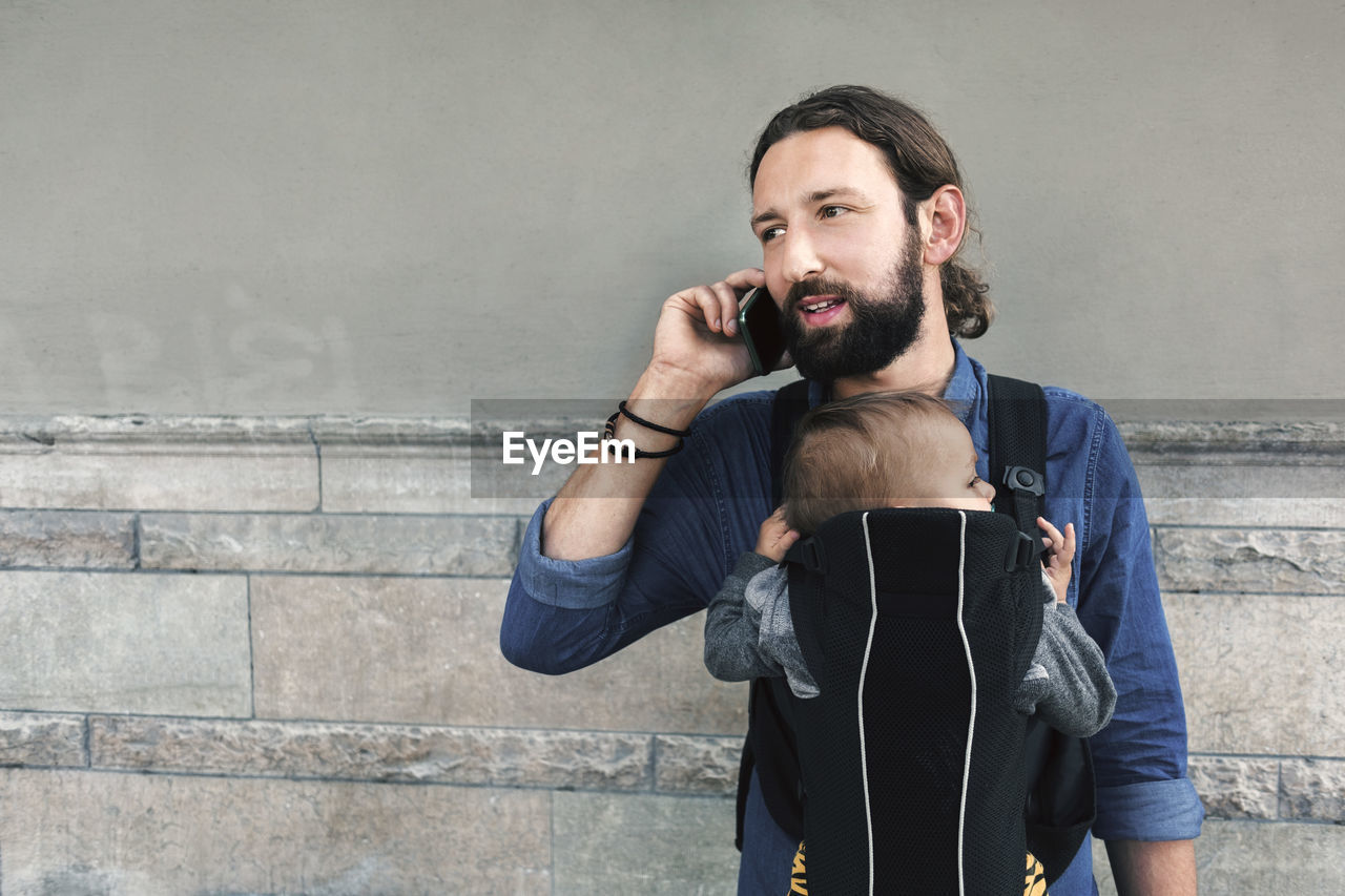 Mid adult man using mobile phone while carrying son in baby carrier against wall