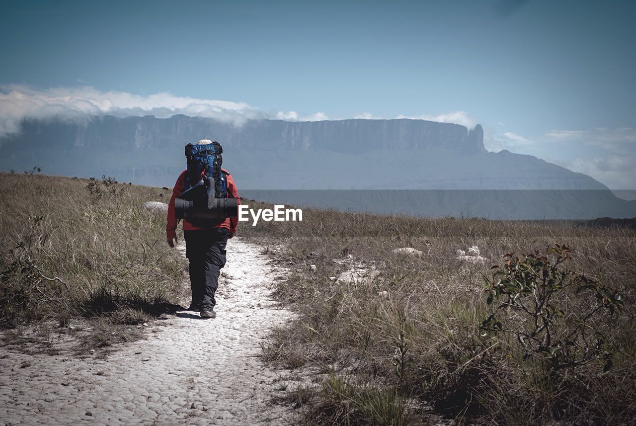 Rear view of hiker walking on dirt road amidst field towards mountains