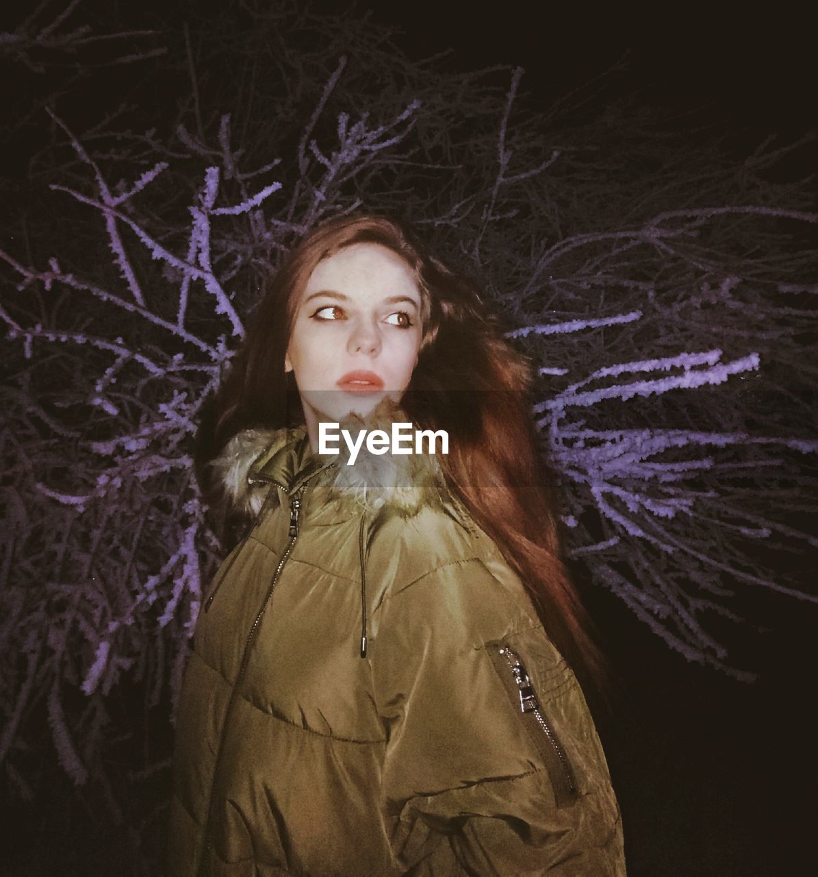 Beautiful young woman standing against tree at night during winter