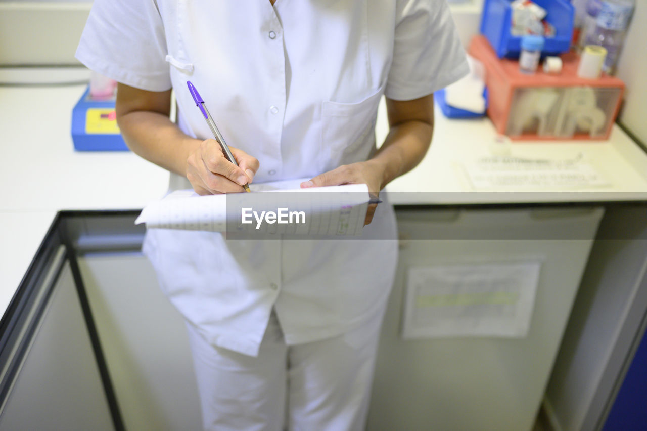 Woman in medical uniform writing on clipboard while standing in office during shift in children hospital