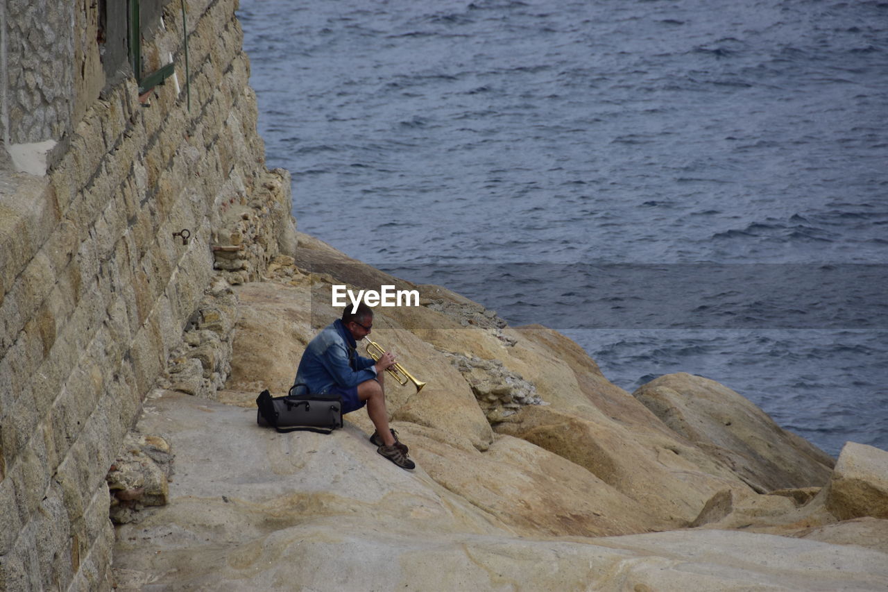 REAR VIEW OF MAN SITTING ON ROCK BY SEA