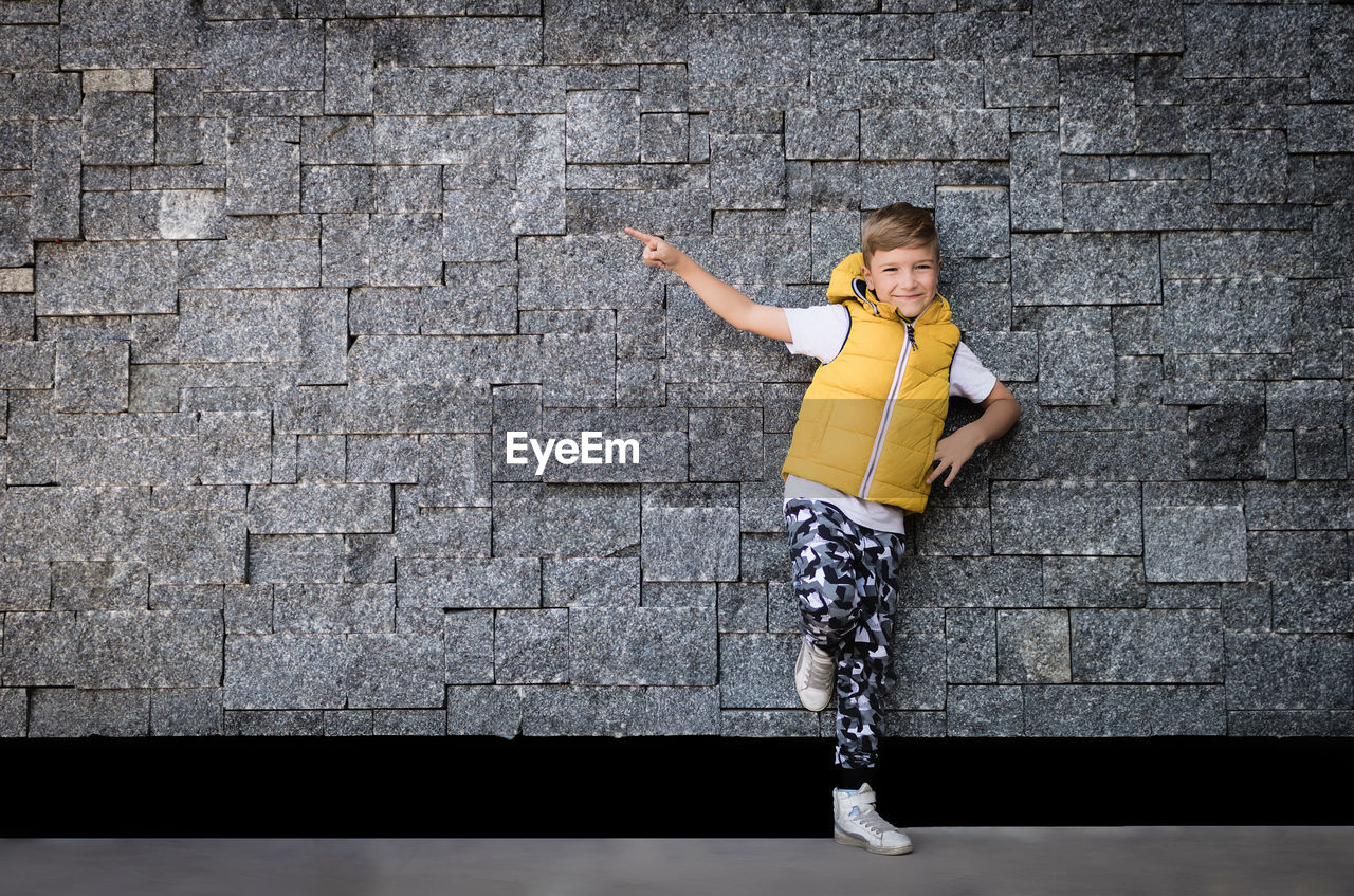 Smiling kid leaning on a brick wall and pointing at it. copy space.