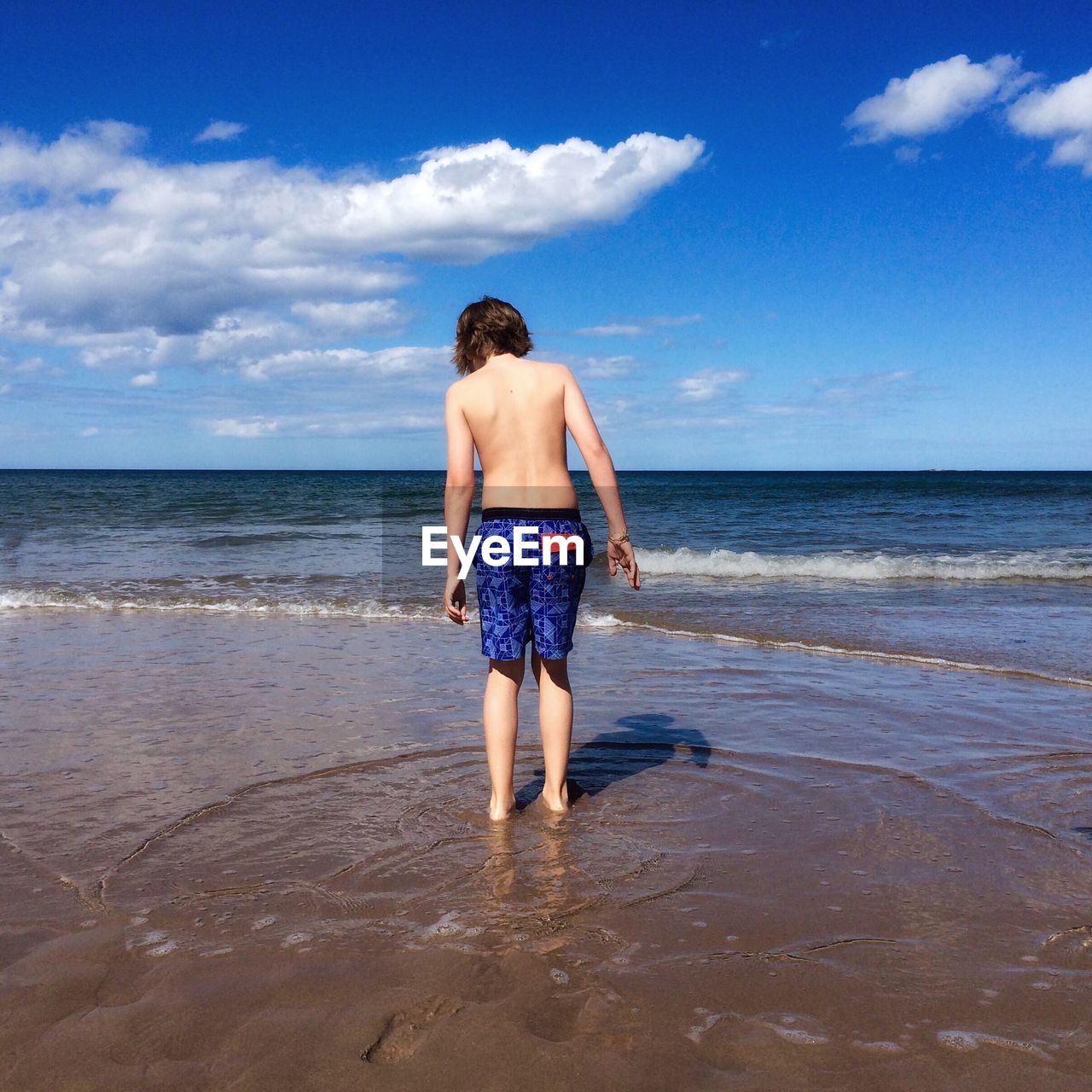 Rear view of shirtless boy standing on sea shore against sky