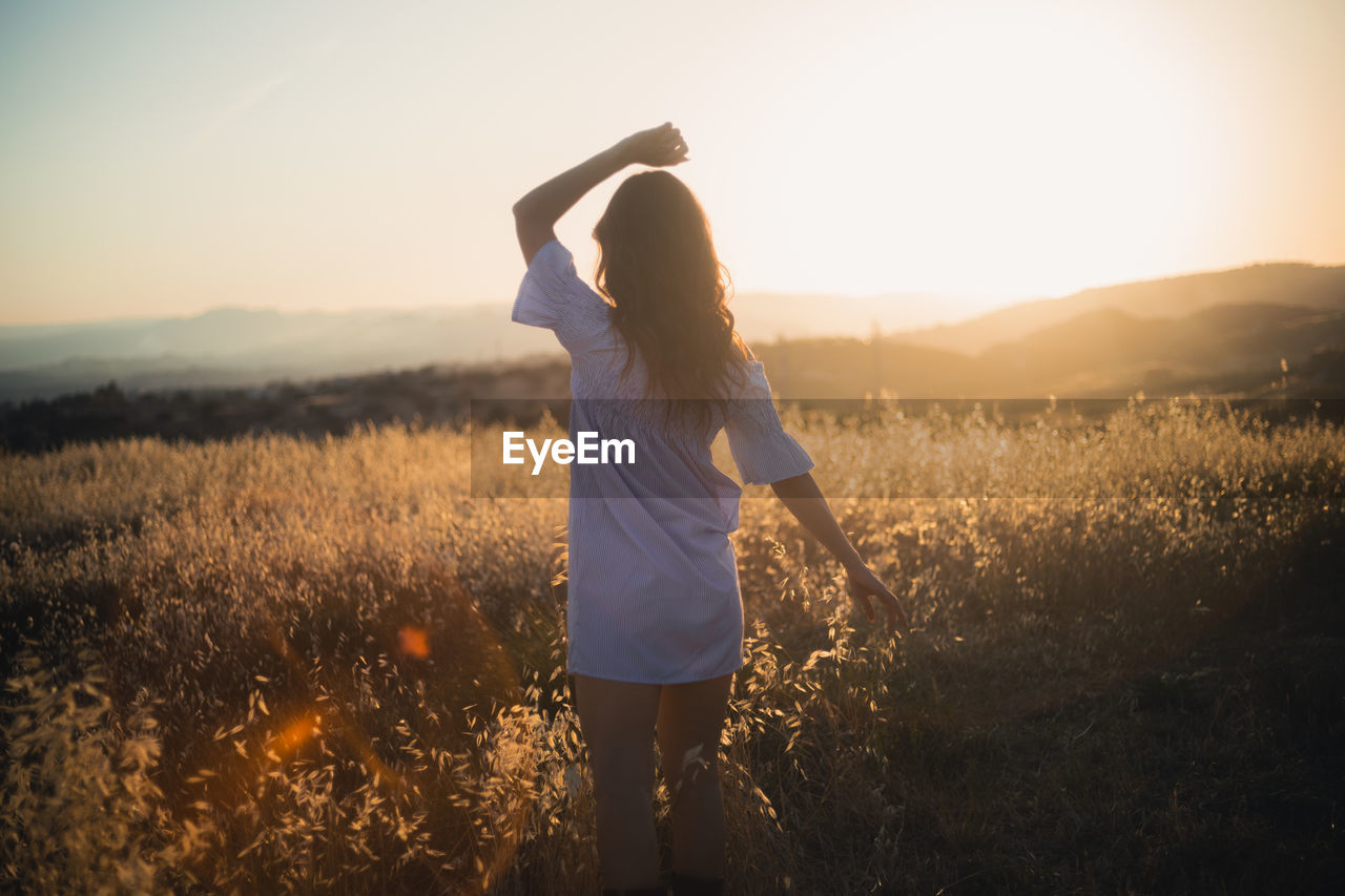 rear view of young woman standing on field against sky during sunset