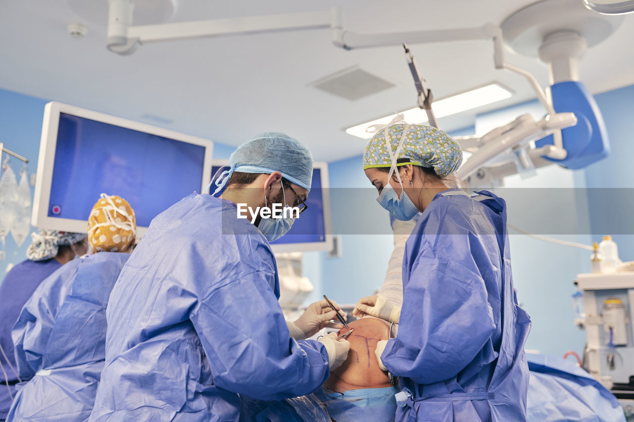 Doctors wearing face mask operating surgery while standing with colleague in background at operation room