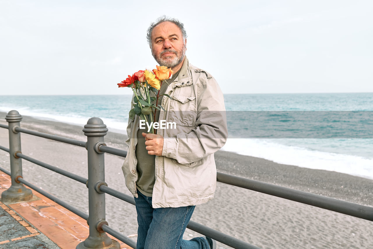 Happy smiling mature bearded man with flower bouquet waiting for girlfriend on seaside promenade.