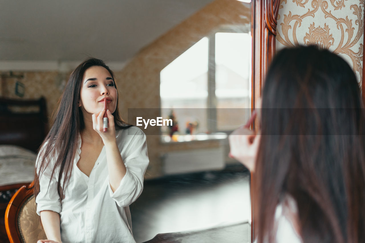 Beautiful woman applying lipstick in front of mirror at home