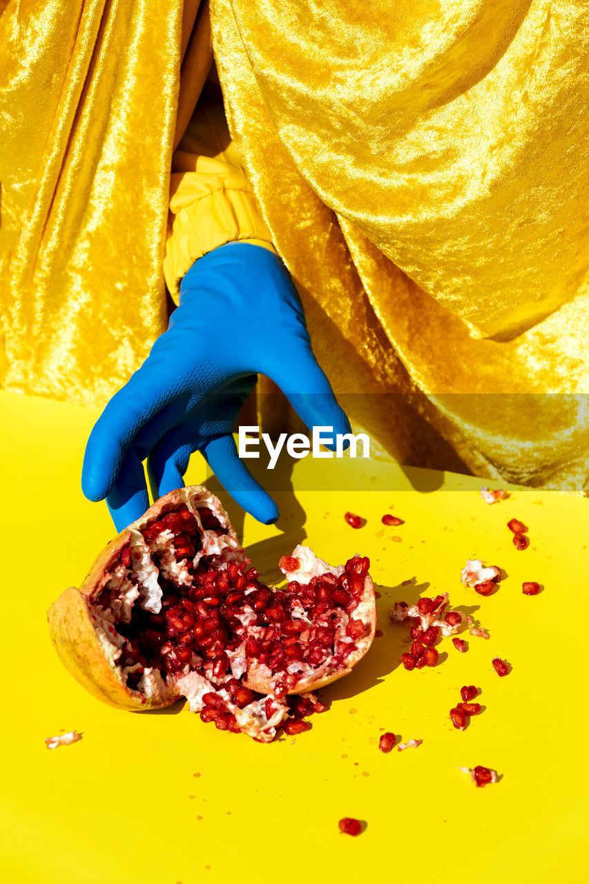 Crop anonymous person in latex glove reaching out hand to smashed ripe pomegranate with red seeds from behind shiny curtain on yellow surface in studio person