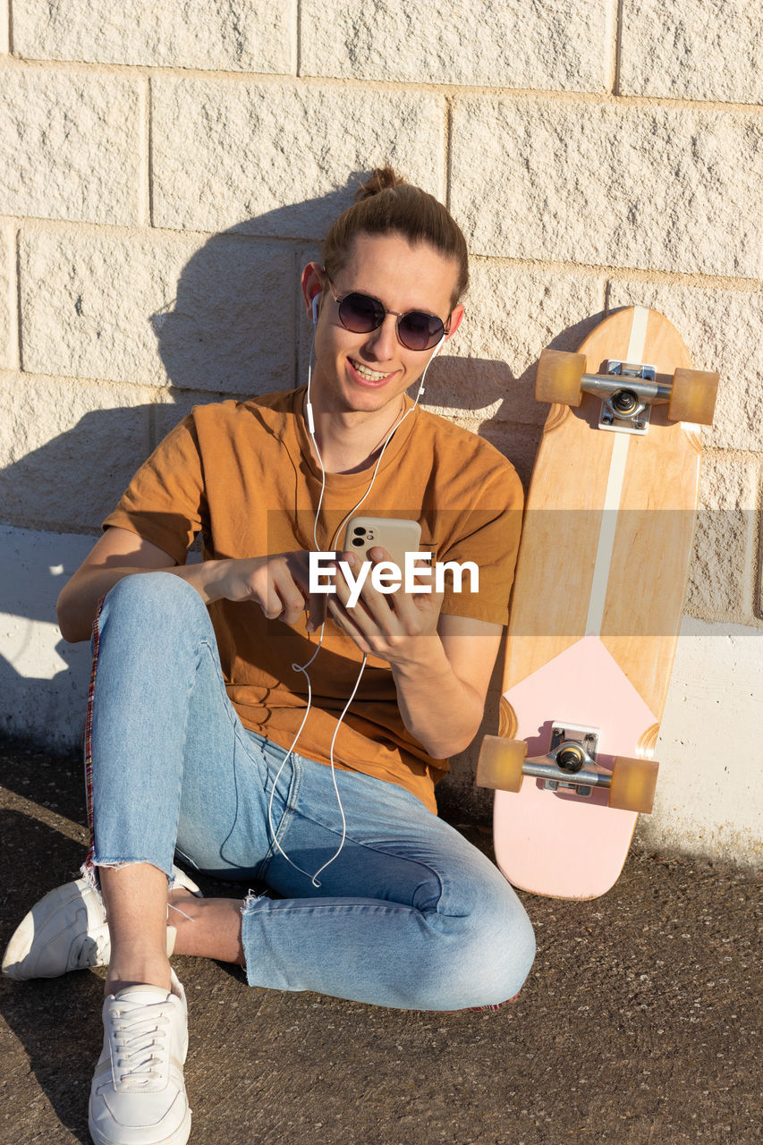 Smiling young man with surf skate on the side sitting on the floor chatting on smartphone having fun