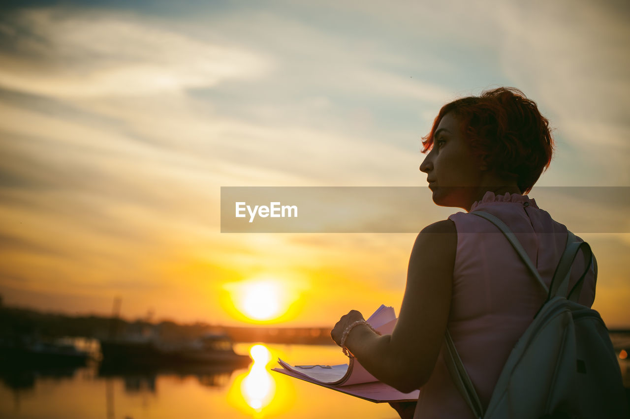 Rear view of mid adult woman holding papers while standing by river against sky during sunset