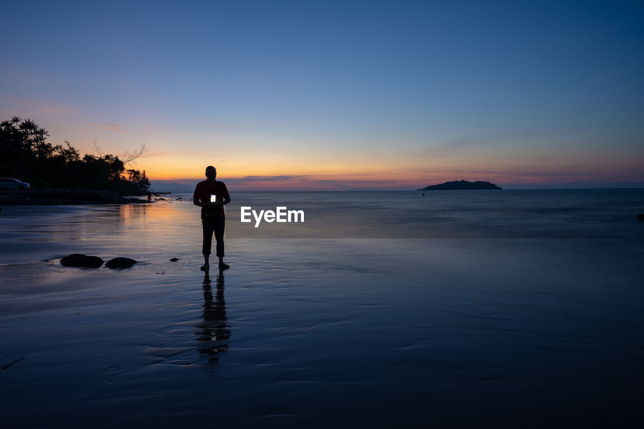 Silhouette man holding camera while standing at beach against sky during sunset