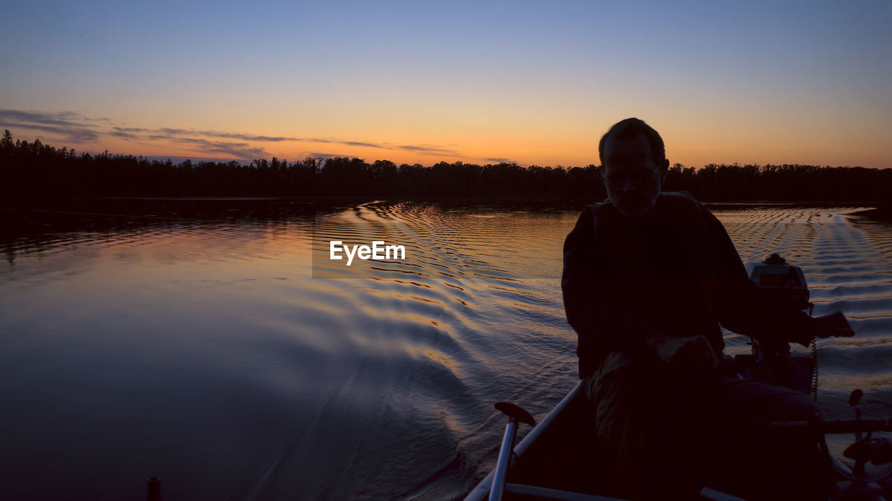 SILHOUETTE MAN SITTING ON BOAT IN LAKE AGAINST SKY DURING SUNSET