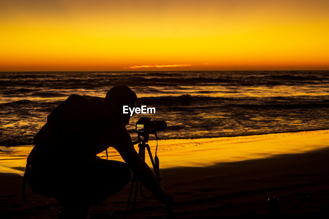 Silhouette man photographing at beach against sky during sunset