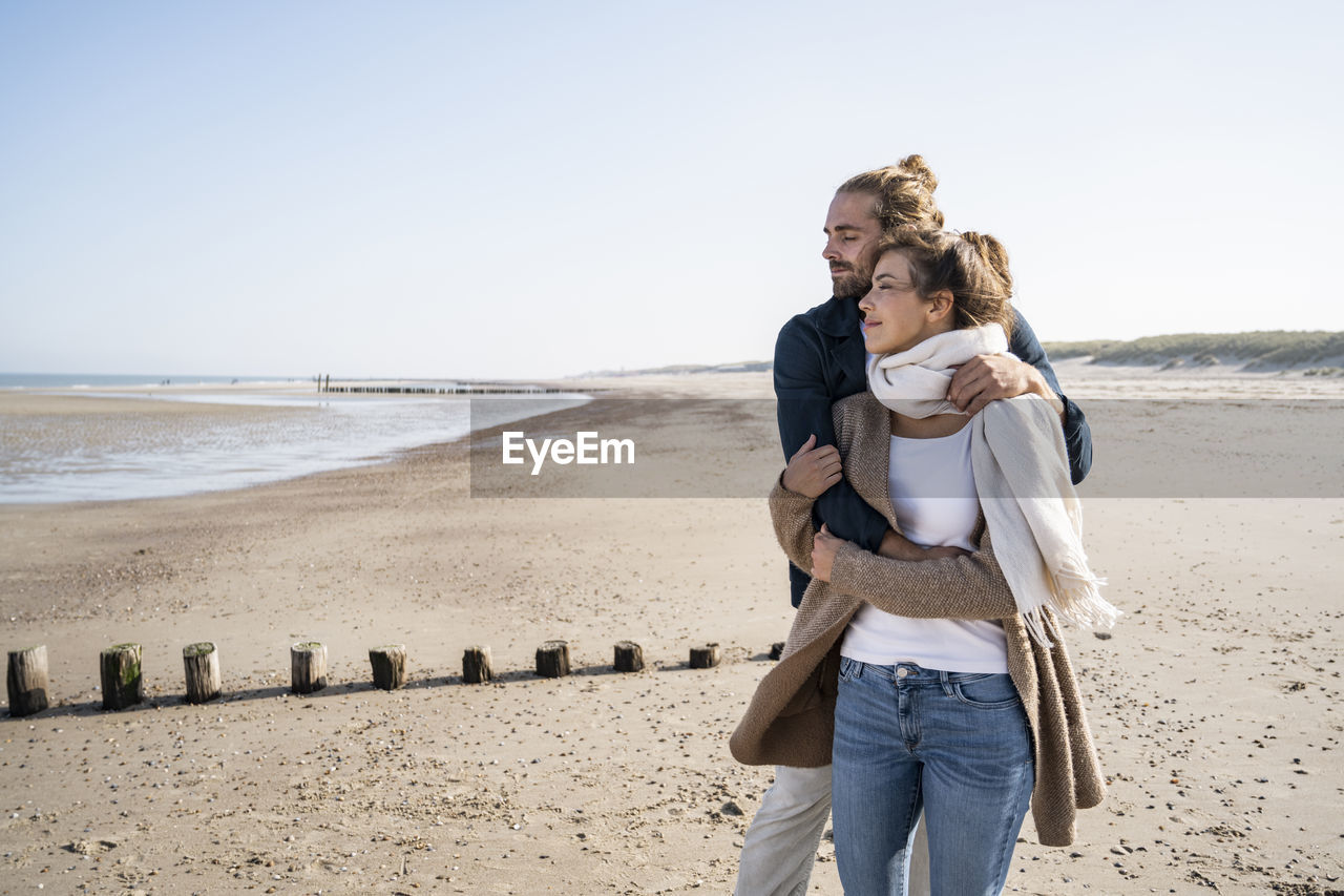 Young couple with eyes closed embracing while standing at beach against clear sky