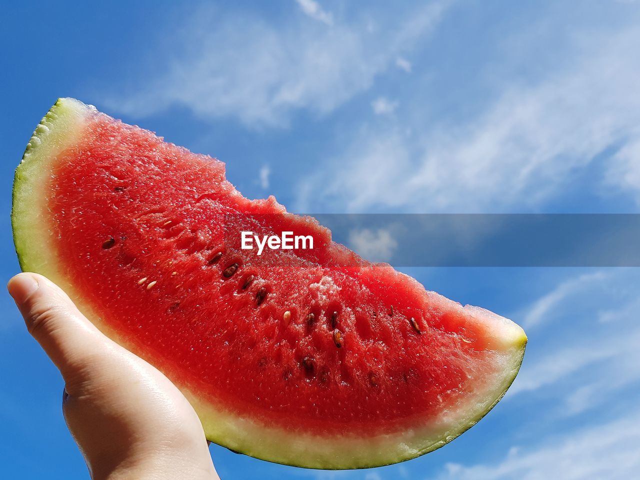 Cropped hand holding watermelon slice against blue sky