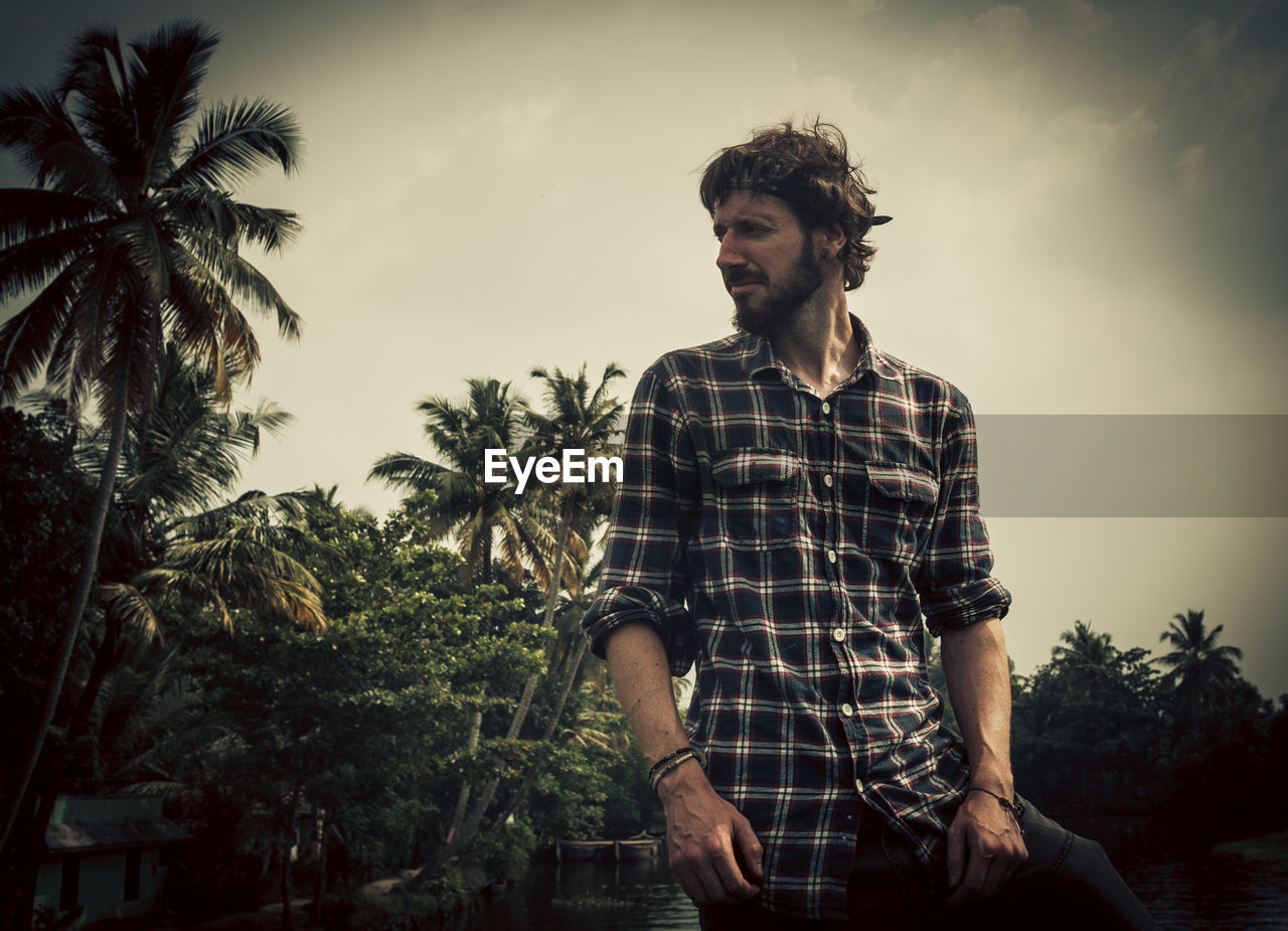 Young man looking away while standing against sky
