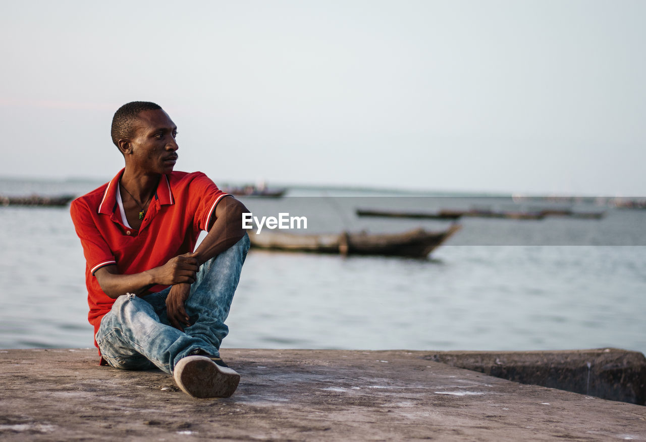 YOUNG MAN SITTING ON SHORE AGAINST SKY