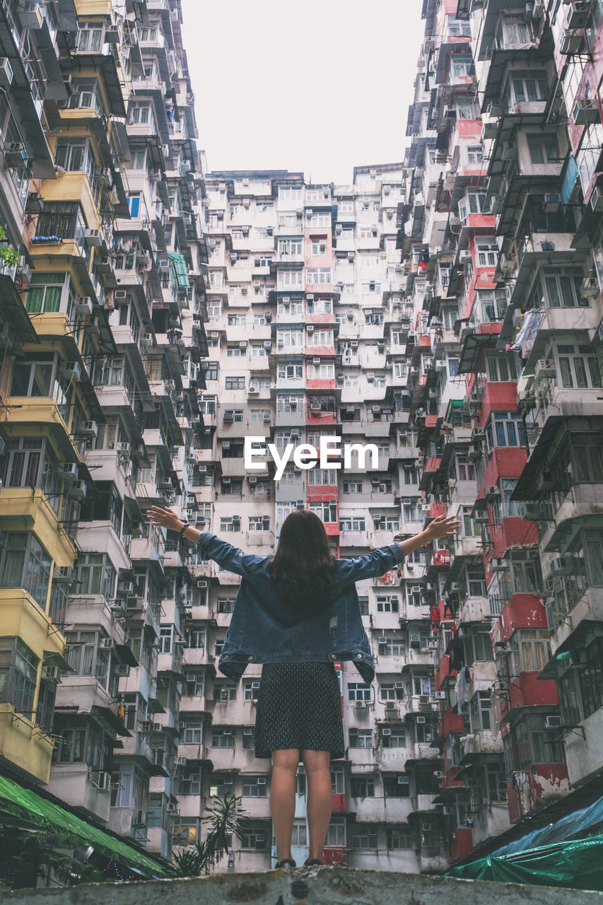 REAR VIEW OF WOMAN STANDING BY BUILDINGS IN CITY AGAINST SKY