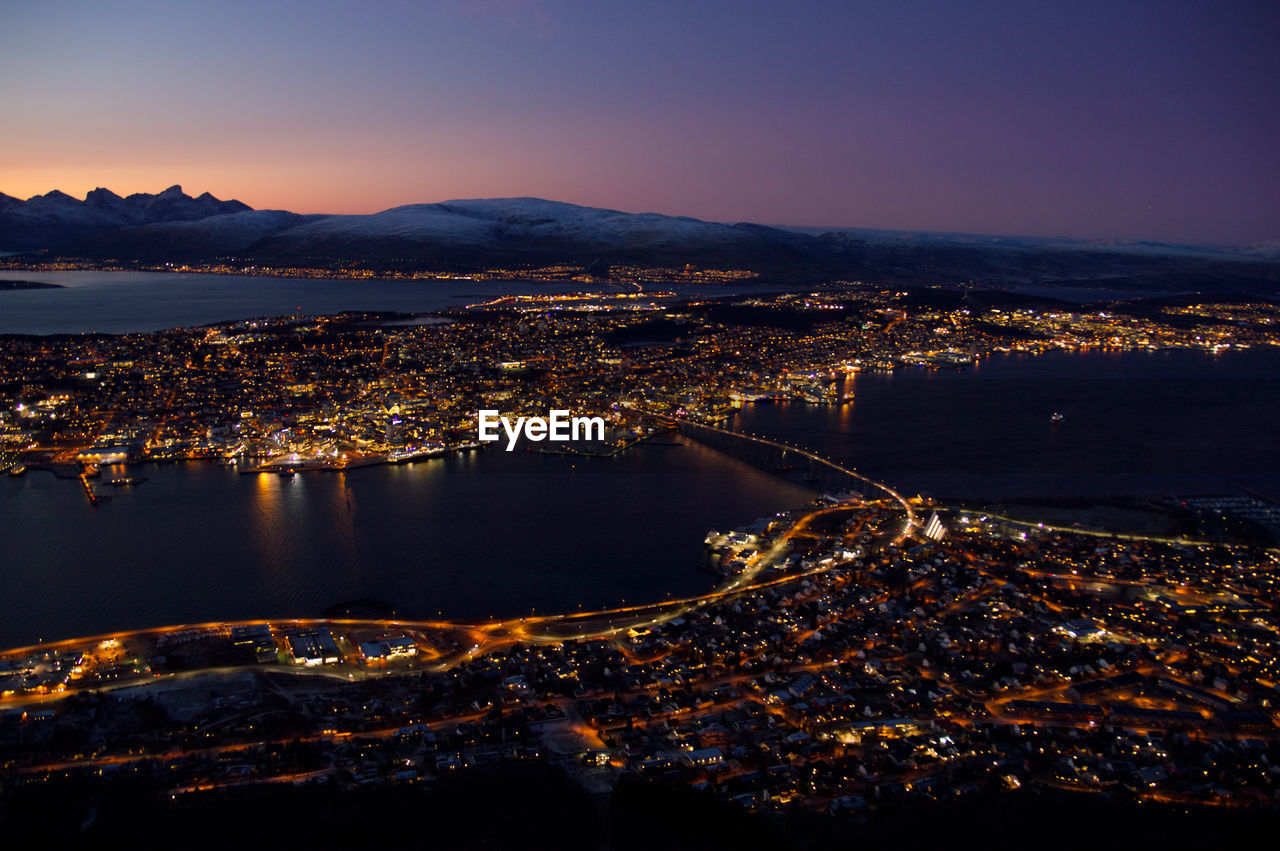 Aerial view of illuminated city by sea against sky at sunset