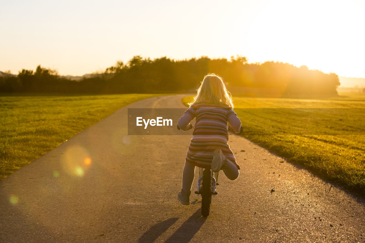 Rear view of girl riding bicycle on road amidst field during sunset