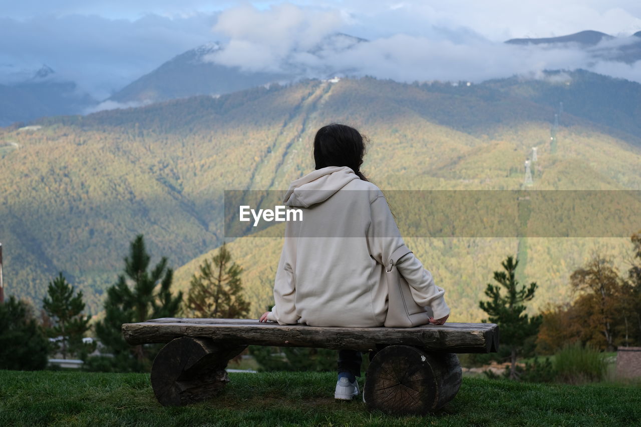 REAR VIEW OF MAN SITTING ON LANDSCAPE AGAINST MOUNTAIN RANGE