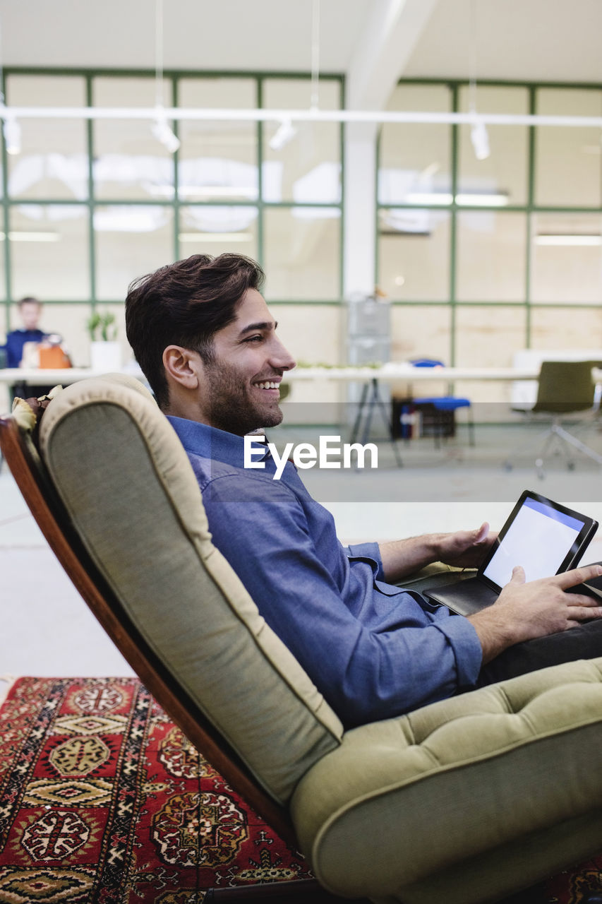 Side view of happy male blogger holding digital tablet while sitting on armchair in office