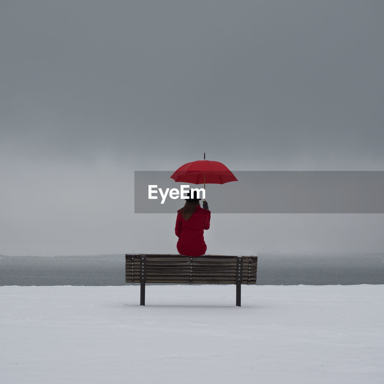 Woman with umbrella sitting on bench at snow covered field