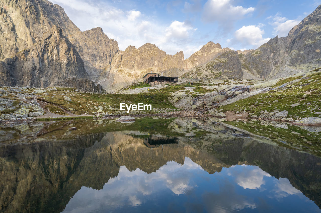 Alpine hut surrounded by mountains reflected in foreground tarn during sunrise, slovakia, europe