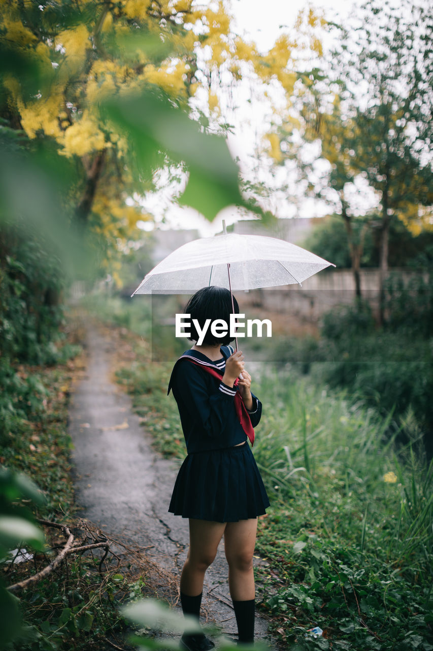 Full length of woman standing on footpath with umbrella during rainy season