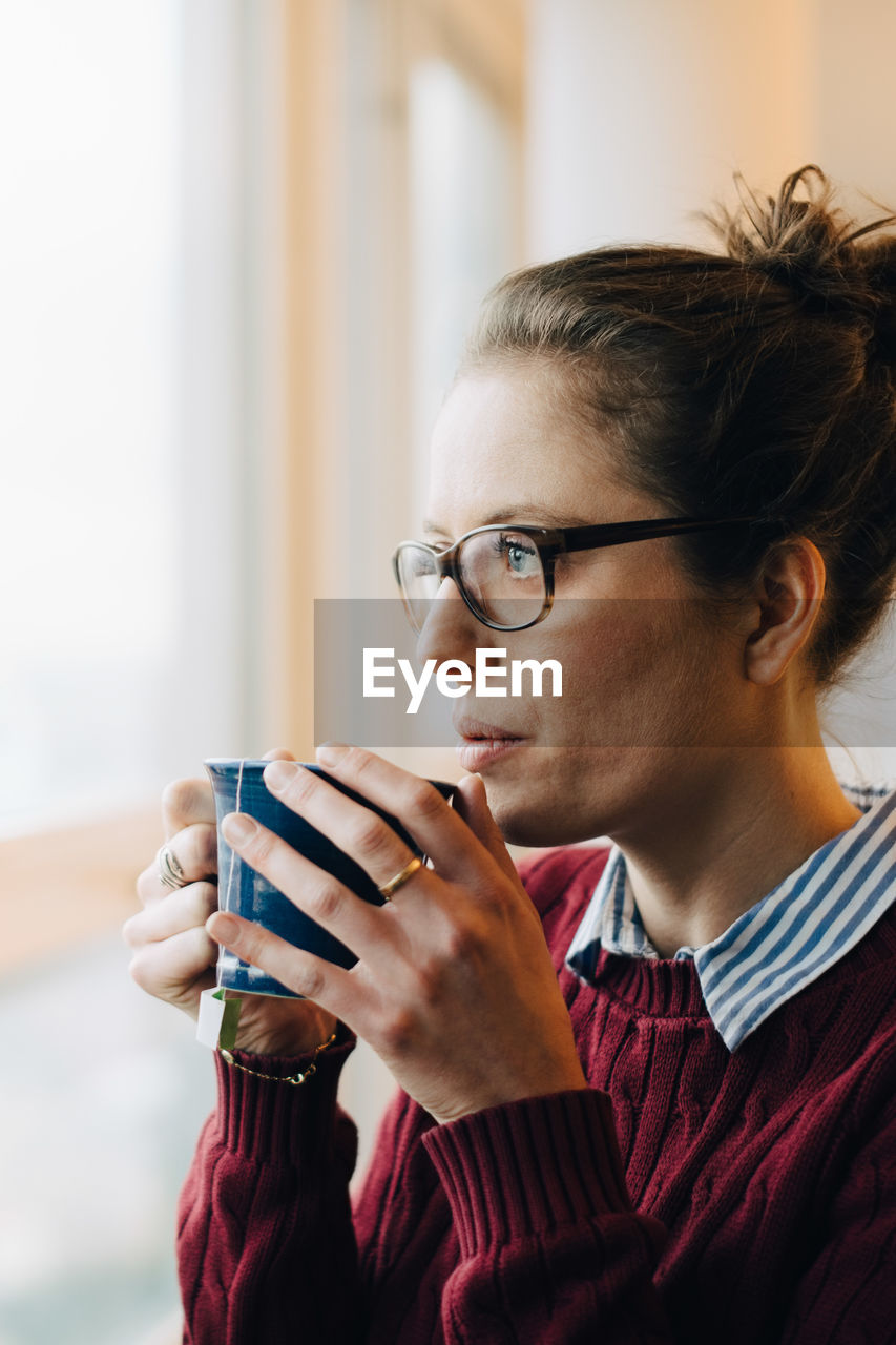 Thoughtful businesswoman drinking coffee while looking through window at creative office