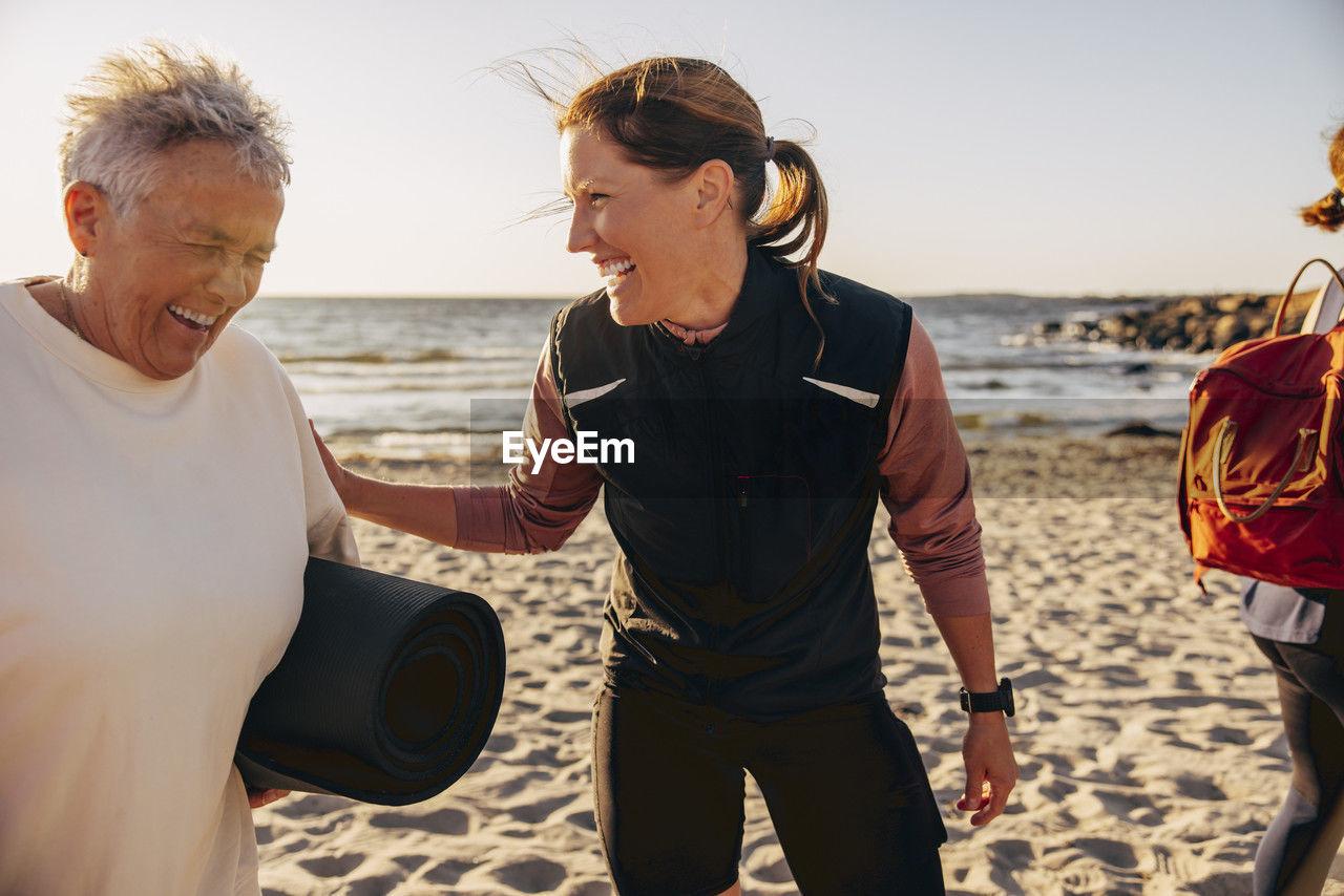Happy female coach laughing with senior woman holding exercise mat at beach