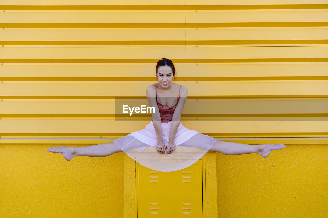 Young woman doing splits while ballet dancing on seat against yellow wall