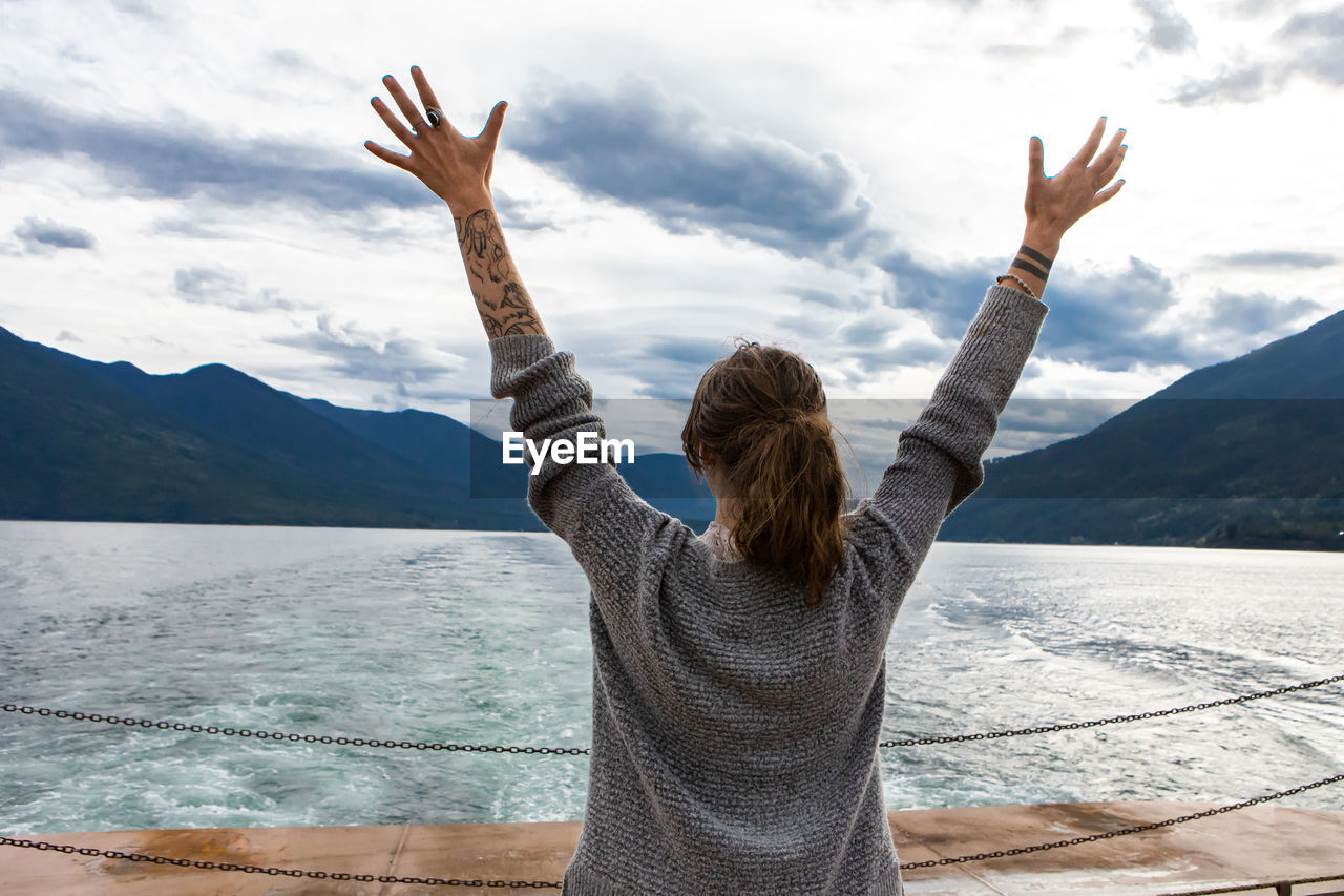 REAR VIEW OF WOMAN WITH ARMS RAISED AGAINST MOUNTAINS