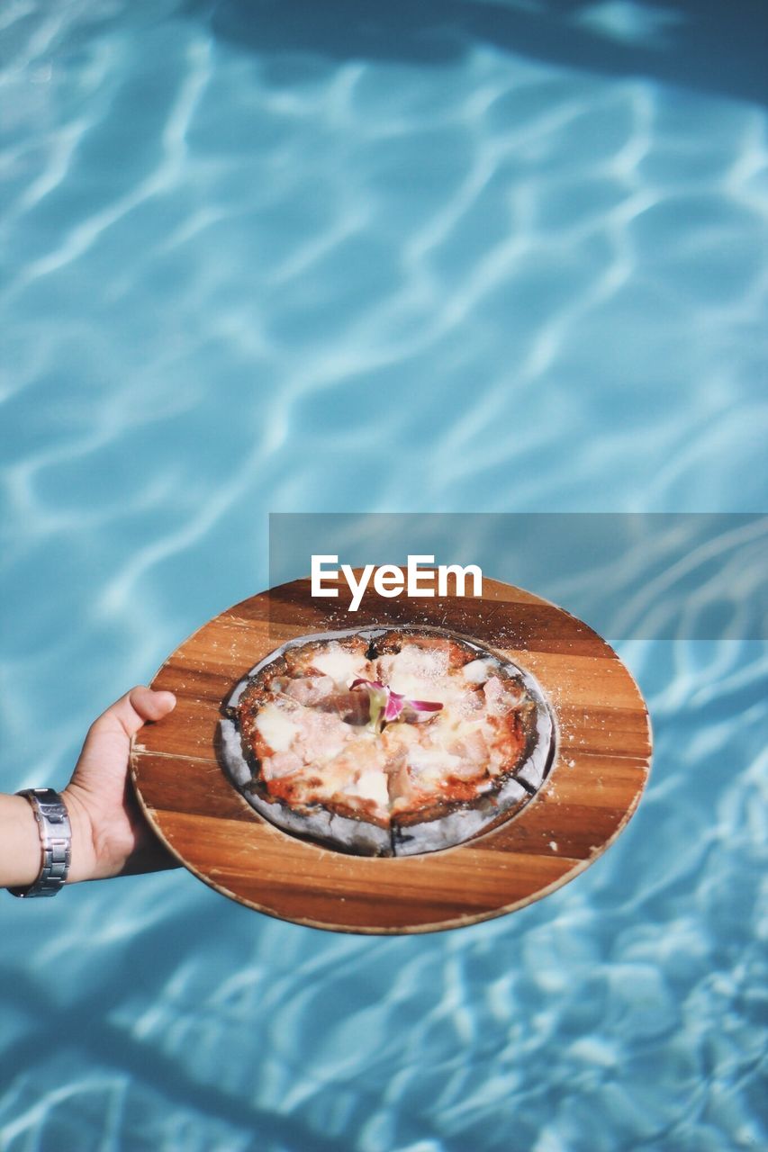 Cropped hand holding pizza in plate over swimming pool