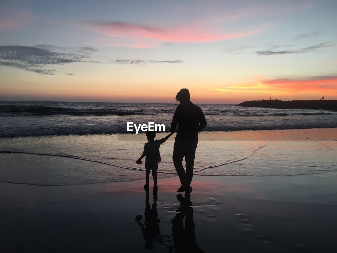 Silhouette father and son walking on beach against sky during sunset