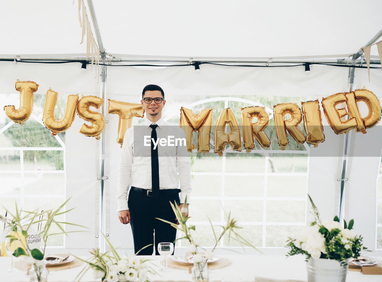 Portrait of smiling bridegroom standing against text in tent