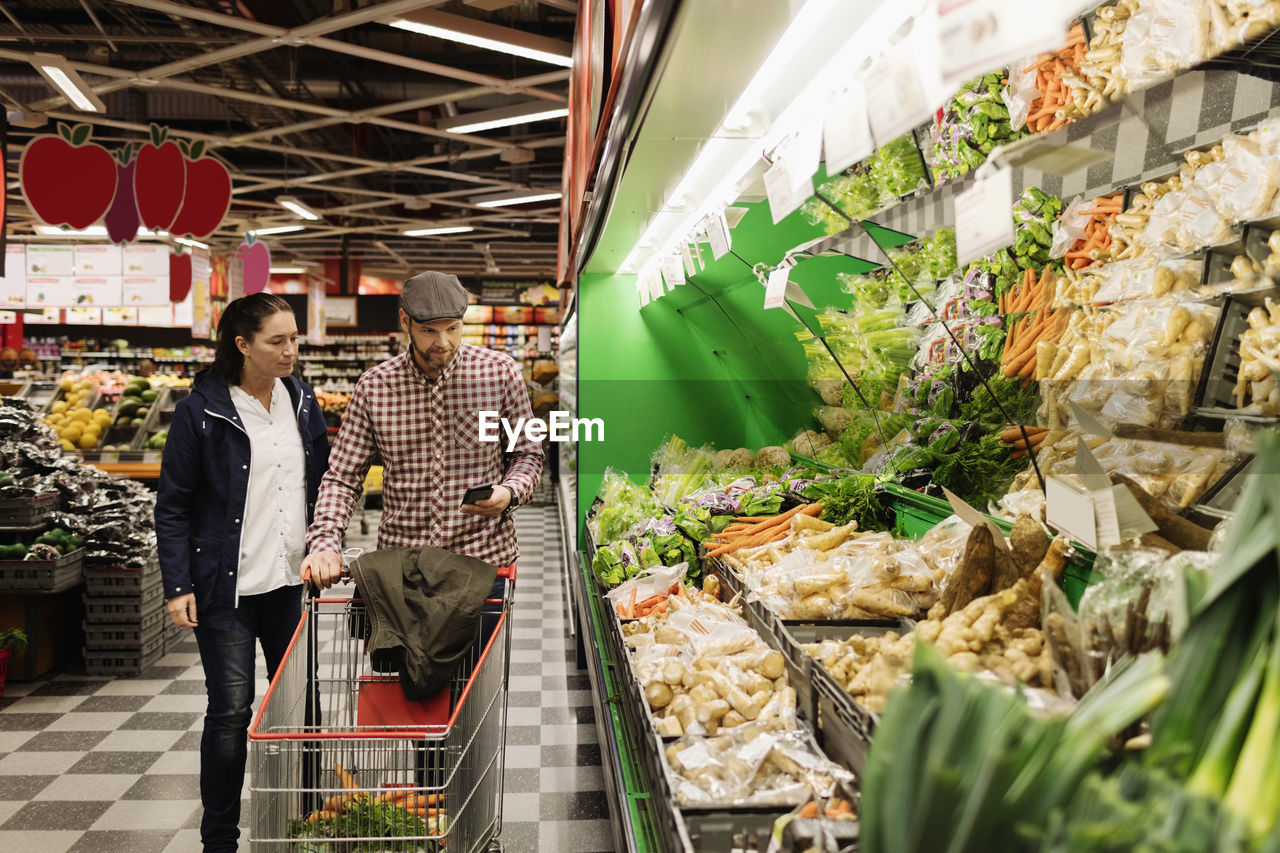 Couple choosing vegetables while using phone in supermarket