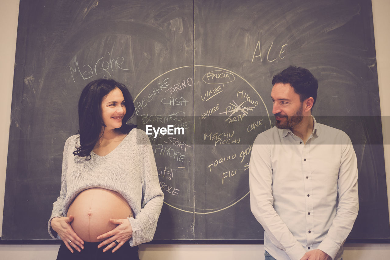 Smiling pregnant woman looking at man against blackboard