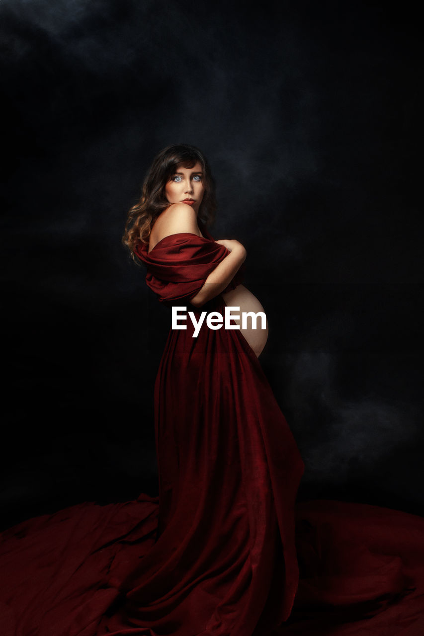 Rear view of pregnant woman staying against black background with red dress