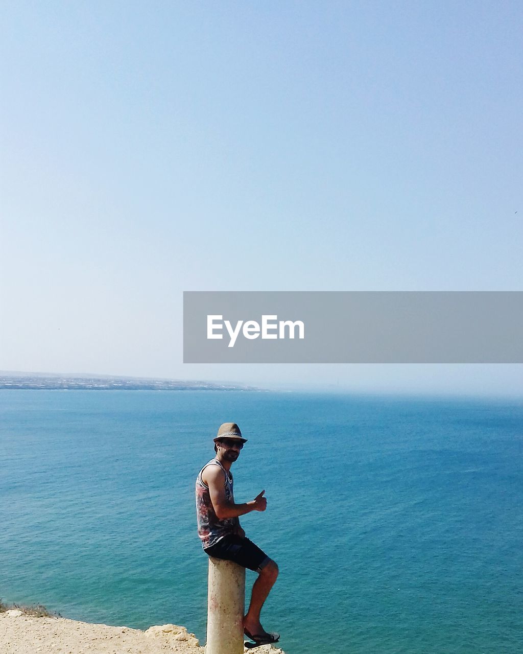 Side view of man sitting on bollard by sea against clear sky